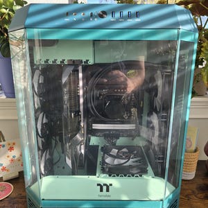 K&ouml;nnte beinhalten: Ein t&uuml;rkis- und mintfarbenes Computergeh&auml;use mit einer transparenten Glasscheibe, das interne Komponenten wie eine Grafikkarte und L&uuml;fter zeigt. Das Geh&auml;use hat ein modernes, eckiges Design mit dem Thermaltake-Logo.
