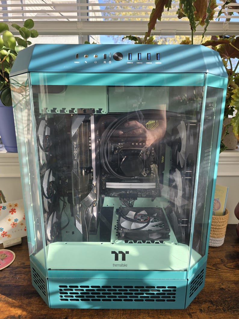 K&ouml;nnte beinhalten: Ein t&uuml;rkis- und mintfarbenes Computergeh&auml;use mit einer transparenten Glasscheibe, das interne Komponenten wie eine Grafikkarte und L&uuml;fter zeigt. Das Geh&auml;use hat ein modernes, eckiges Design mit dem Thermaltake-Logo.
