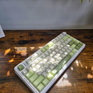 May include: A white mechanical keyboard with light green and white keycaps. The keyboard has a compact layout with a volume knob and is set on a wooden surface. The keycaps are a mix of light green and white, creating a clean and modern aesthetic.