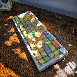 May include: A pastel-colored mechanical keyboard with a white frame and a silver knob. The keycaps are a mix of soft colors, including yellow, green, blue, pink, and orange. The keyboard sits on a dark wooden surface.