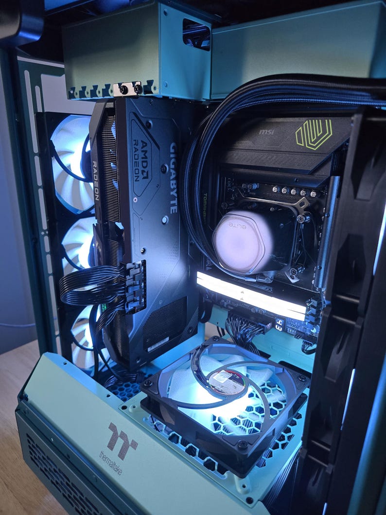 K&ouml;nnte beinhalten: Ein individuell gebautes Computergeh&auml;use in T&uuml;rkis, das interne Komponenten zeigt. Das Bild zeigt eine Grafikkarte mit AMD Radeon- und Gigabyte-Branding, zusammen mit mehreren L&uuml;ftern und LED-Beleuchtung in Blau und Wei&szlig;. Das Geh&auml;use ist offen.