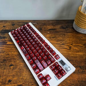 May include: A white mechanical keyboard with red translucent keycaps. The keyboard has a small screen and a silver dial on the right side. The keys are arranged in a standard layout.