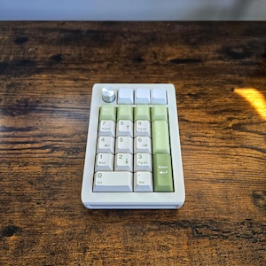 May include: A compact, off-white and green mechanical number pad with a silver knob. The keys are arranged in a standard numeric layout, with white and green keycaps. The number pad sits on a dark wooden surface.