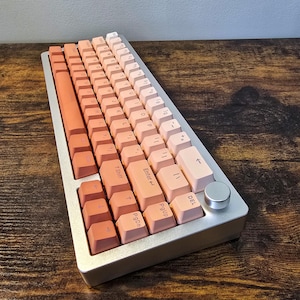 May include: A mechanical keyboard with a gradient color scheme, transitioning from coral to light pink. The keyboard has a silver frame and a round, silver knob. The keycaps are arranged in a standard layout.