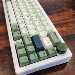 May include: A white mechanical keyboard with green and off-white keycaps. The keyboard has a volume knob and a variety of keys, including function keys, arrow keys, and a backspace key. The keyboard sits on a wooden surface.