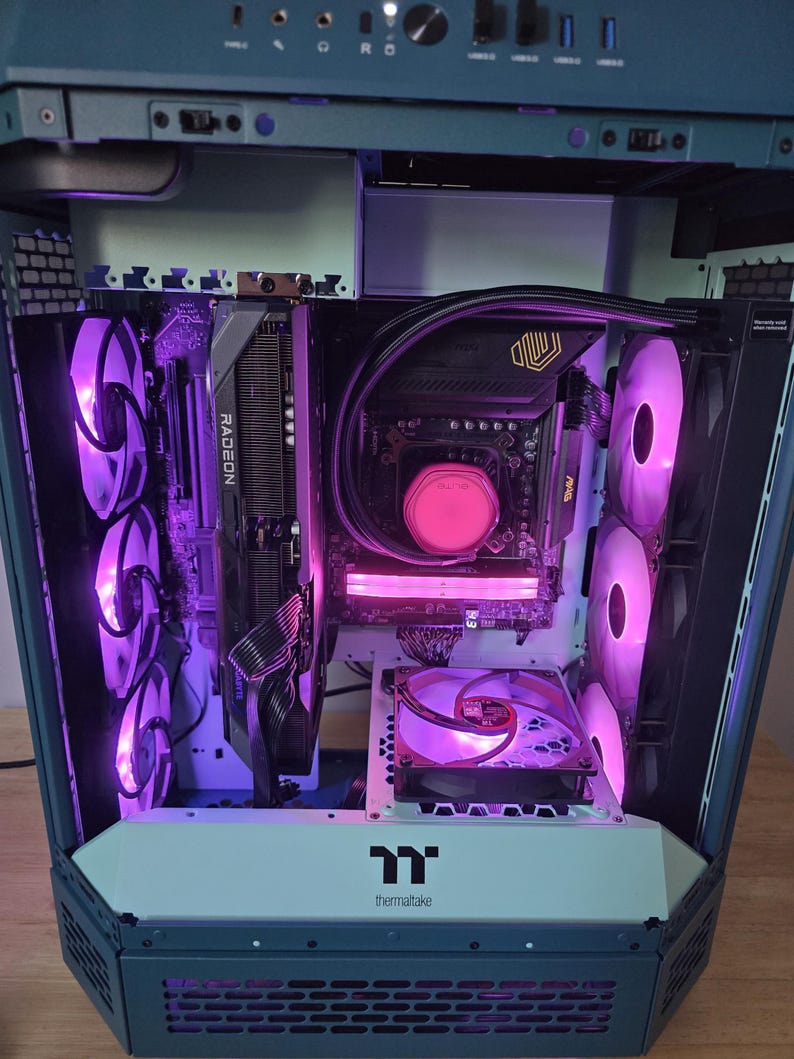 K&ouml;nnte beinhalten: Ein Computergeh&auml;use mit internen Komponenten, die von violetten Lichtern beleuchtet werden. Das Geh&auml;use hat ein Thermaltake-Logo. Zu den Komponenten geh&ouml;ren eine Radeon-Grafikkarte und mehrere L&uuml;fter.