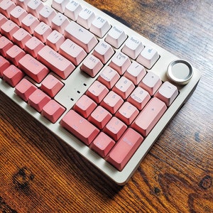 May include: A mechanical keyboard with a pink and white gradient keycap design. The keyboard has a gold-coloured frame and a volume control knob. The keys are arranged in a standard layout, with function keys and a number pad.