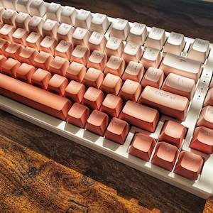May include: A mechanical keyboard with a gradient color scheme, transitioning from white to coral. The keys are arranged in a standard layout with function keys and a gold-colored volume knob. The keyboard sits on a wooden surface.