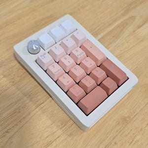 May include: A white and pink mechanical number pad with a silver dial. The keycaps are a gradient of pink hues, with white keys at the top. The keys are labeled with numbers and symbols.