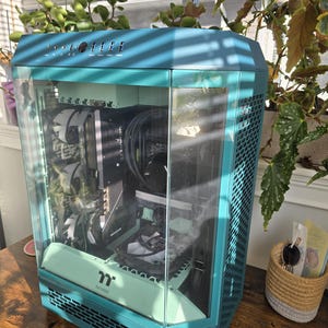 K&ouml;nnte beinhalten: Ein t&uuml;rkisfarbenes Computergeh&auml;use mit Glasfront, das interne Komponenten zeigt. Das Geh&auml;use hat ein sechseckiges Design und einen mintgr&uuml;nen Akzent. Das Geh&auml;use steht auf einer Holzoberfl&auml;che, mit Pflanzen im Hintergrund.