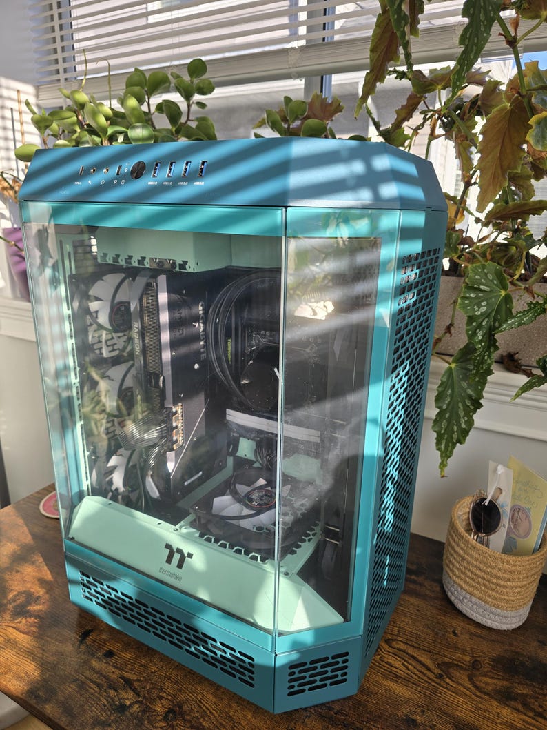 K&ouml;nnte beinhalten: Ein t&uuml;rkisfarbenes Computergeh&auml;use mit Glasfront, das interne Komponenten zeigt. Das Geh&auml;use hat ein sechseckiges Design und einen mintgr&uuml;nen Akzent. Das Geh&auml;use steht auf einer Holzoberfl&auml;che, mit Pflanzen im Hintergrund.