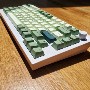 May include: A white mechanical keyboard with green and white keycaps. The keycaps have varying shades of green, creating a gradient effect. A silver knob is on the right side. The keyboard sits on a wooden surface.