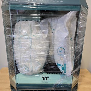 K&ouml;nnte beinhalten: Ein teal-farbenes und schwarzes Computergeh&auml;use, in transparente Folie gewickelt, mit dem Thermaltake-Logo. Das Geh&auml;use hat ein geometrisches Design und enth&auml;lt mehrere USB-Anschl&uuml;sse und Kn&ouml;pfe auf der Oberseite. Der Innenraum ist mit Schutzverpackungen gef&uuml;llt.