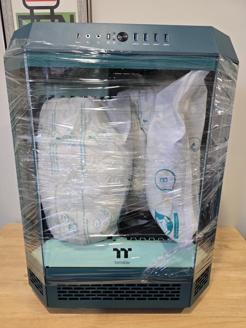 K&ouml;nnte beinhalten: Ein teal-farbenes und schwarzes Computergeh&auml;use, in transparente Folie gewickelt, mit dem Thermaltake-Logo. Das Geh&auml;use hat ein geometrisches Design und enth&auml;lt mehrere USB-Anschl&uuml;sse und Kn&ouml;pfe auf der Oberseite. Der Innenraum ist mit Schutzverpackungen gef&uuml;llt.