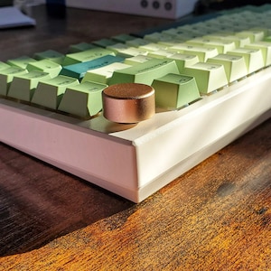 May include: A mechanical keyboard with light green keycaps and a rose gold knob. The keyboard has a white base and is set on a wooden surface. The keycaps have a gradient of green hues.