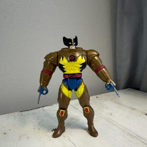 May include: A brown and yellow action figure of Wolverine wearing a brown and silver armoured suit. The figure has a black head with a yellow mask and a red X on the chest.