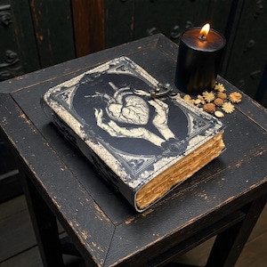 May include: A gothic-style journal with a black cover featuring a detailed illustration of a heart held in hands. The journal has a metal clasp and aged-looking pages. A lit black candle and dried flowers sit nearby on a distressed black table.