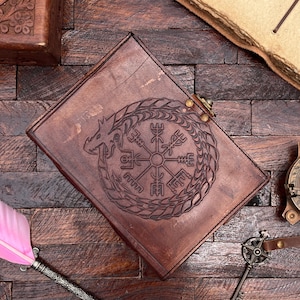May include: A brown leather journal with a compass and dragon design embossed on the cover. The journal has a brass clasp closure.