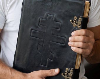 May include: A black leather-bound book with a cross design embossed on the cover. The book has a clasp closure and aged-looking pages. The cover is held by a person wearing a white t-shirt.
