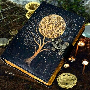 Puede incluir: Un diario de cuero negro con un dise&ntilde;o de &aacute;rbol y luna dorados. El diario tiene un cierre de metal y est&aacute; decorado con estrellas doradas.