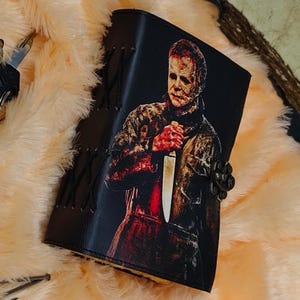 May include: A black leather journal with a graphic of Michael Myers from the Halloween movie series. The journal has a metal clasp and is decorated with stitching.