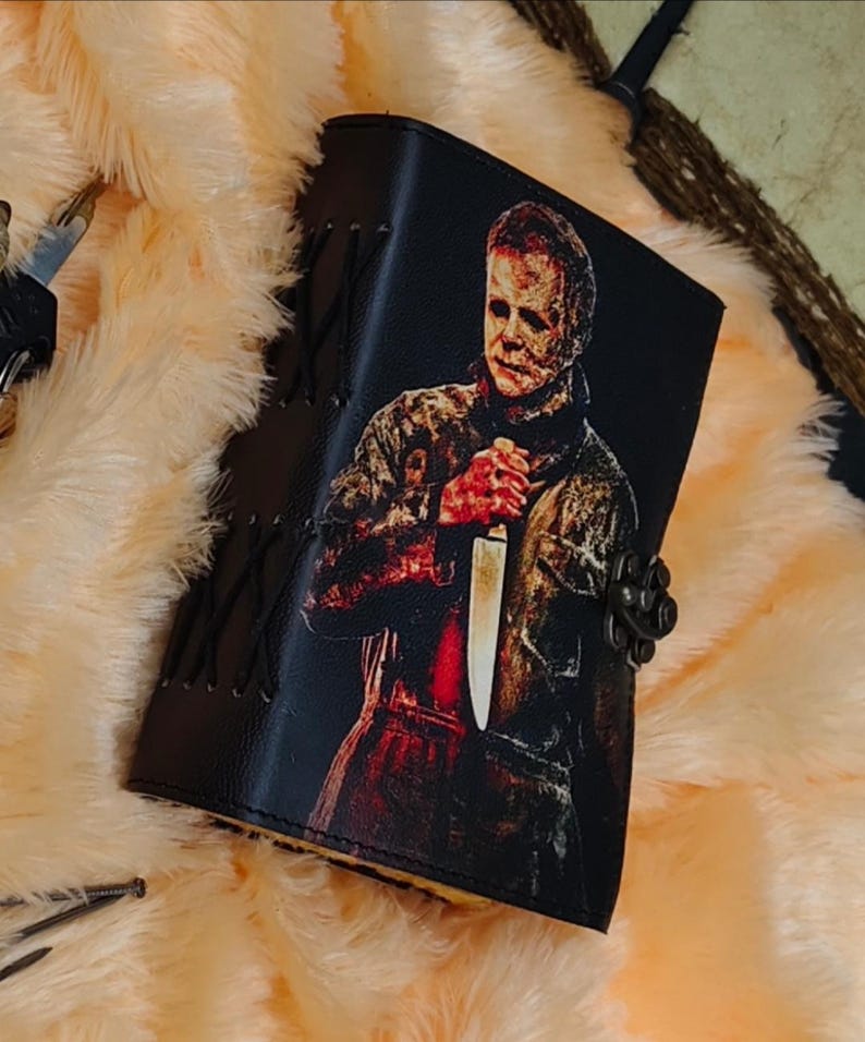 May include: A black leather journal with a graphic of Michael Myers from the Halloween movie series. The journal has a metal clasp and is decorated with stitching.