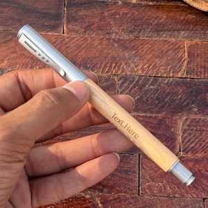 Puede incluir: Un bol&iacute;grafo de madera con una parte superior plateada y una inscripci&oacute;n personalizada que dice "Text Here".