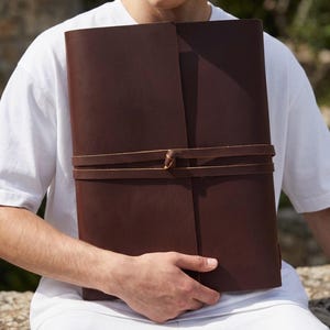 May include: A large, dark brown leather journal with a wrap-around tie closure. The journal is held by a person wearing a white t-shirt and white pants. The leather appears to be smooth and the journal is rectangular in shape.