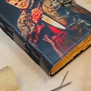May include: A black leather-bound journal with a stitched spine and a metal clasp. The cover features a graphic image of a horror movie character holding a knife.