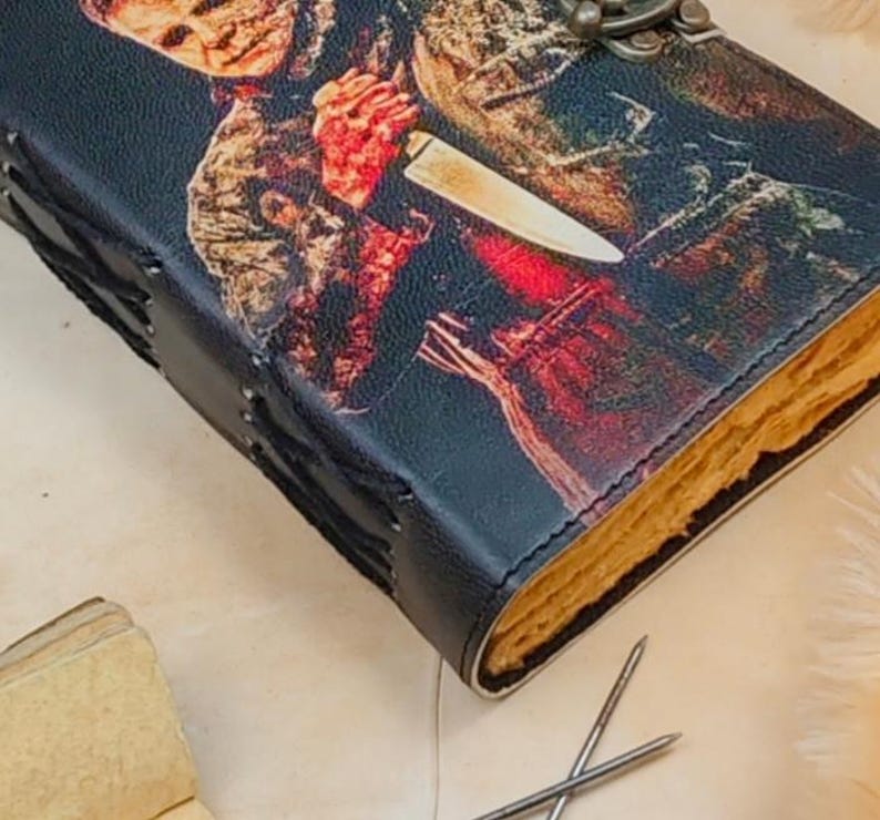 May include: A black leather-bound journal with a stitched spine and a metal clasp. The cover features a graphic image of a horror movie character holding a knife.