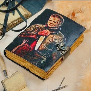 May include: A black leather journal with a colorful illustration of a person holding a knife. The journal has a metal clasp and is closed. The journal is on a light brown surface.