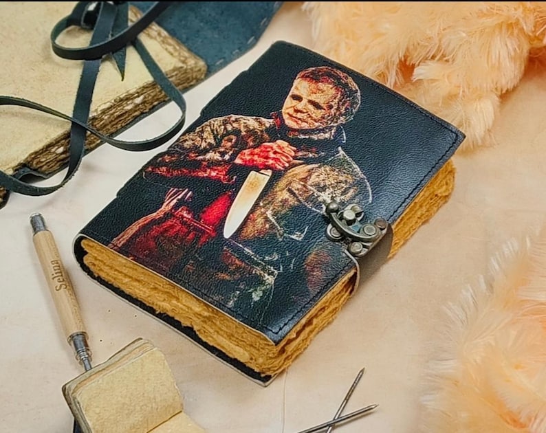May include: A black leather journal with a colorful illustration of a person holding a knife. The journal has a metal clasp and is closed. The journal is on a light brown surface.