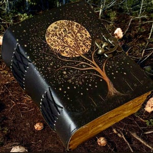 Puede incluir: Un diario de cuero negro con un dise&ntilde;o de &aacute;rbol dorado y una luna en el fondo. El diario tiene un cierre de metal y est&aacute; decorado con estrellas.