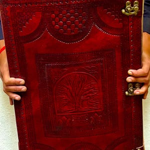 Pode incluir: Um grande di&aacute;rio retangular de couro vermelho com um desenho de &aacute;rvore da vida em relevo na capa. O di&aacute;rio tem fechos cor de lat&atilde;o e uma borda decorativa. O di&aacute;rio &eacute; segurado por uma pessoa.