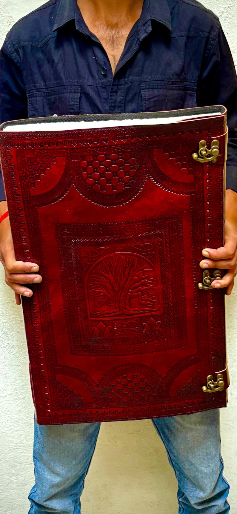 Pode incluir: Um grande di&aacute;rio retangular de couro vermelho com um desenho de &aacute;rvore da vida em relevo na capa. O di&aacute;rio tem fechos cor de lat&atilde;o e uma borda decorativa. O di&aacute;rio &eacute; segurado por uma pessoa.