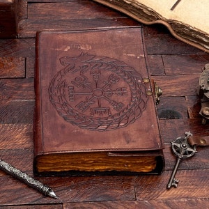 May include: A brown leather journal with a compass and dragon design embossed on the cover. The journal is closed and lying on a wooden surface.