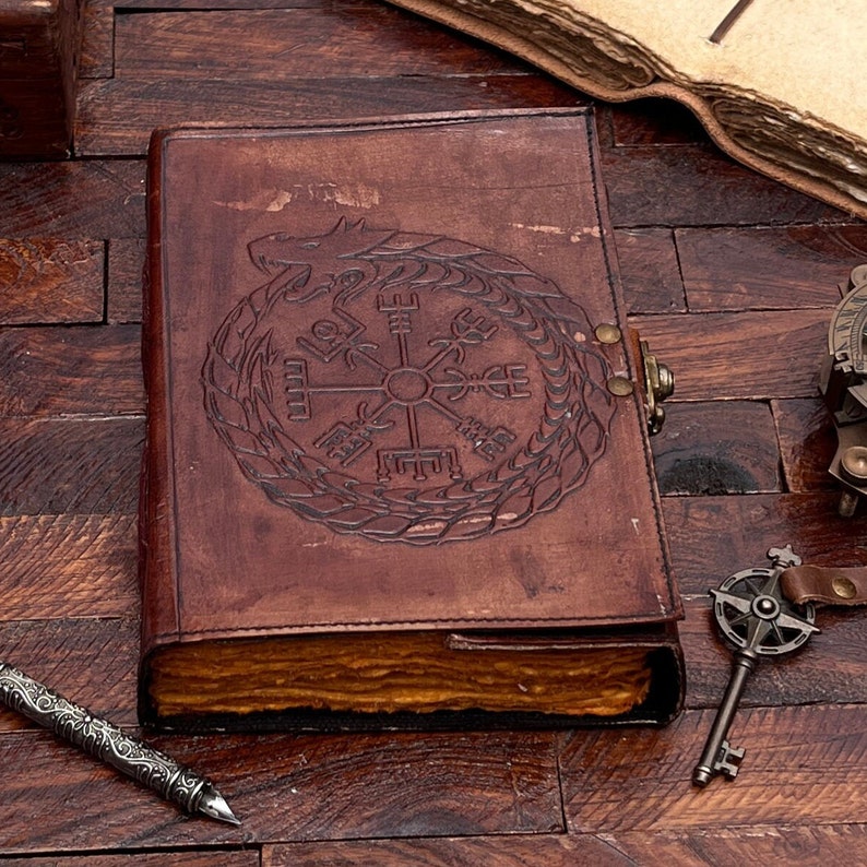 May include: A brown leather journal with a compass and dragon design embossed on the cover. The journal is closed and lying on a wooden surface.