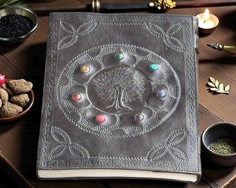 May include: A gray leather journal with an embossed tree of life design. The cover features a circular pattern with colorful gemstone accents. The journal is open, revealing blank pages, and is surrounded by other decorative items.