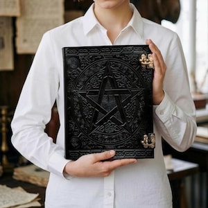 May include: Black leather journal with a raised pentagram design and ornate border. The journal has a clasp closure and is held by a person wearing a white button-down shirt. The journal is a dark color and has a vintage aesthetic.