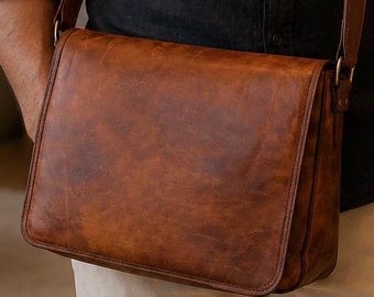 May include: A brown leather messenger bag with a rectangular flap, visible stitching along the edges, and a matching adjustable shoulder strap. The surface of the bag displays a natural, textured grain with subtle variations in the brown color tone.