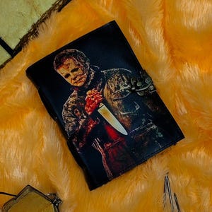 May include: A black leather journal with a graphic of a horror movie character holding a knife. The journal has a metal clasp.