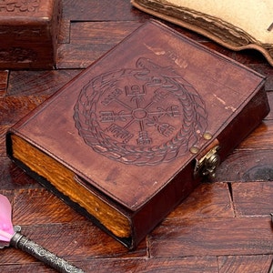 May include: A brown leather journal with a decorative embossed design of a compass surrounded by a serpent. The journal has a brass clasp and a worn, aged look.