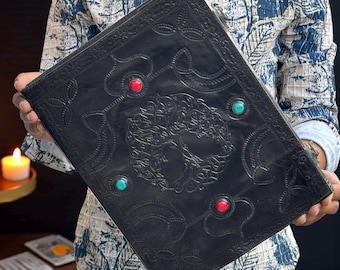 May include: A large, black leather journal with an embossed tree of life design. The cover is adorned with red and turquoise colored stone accents. The journal is held open, showcasing the intricate details of the embossed artwork.