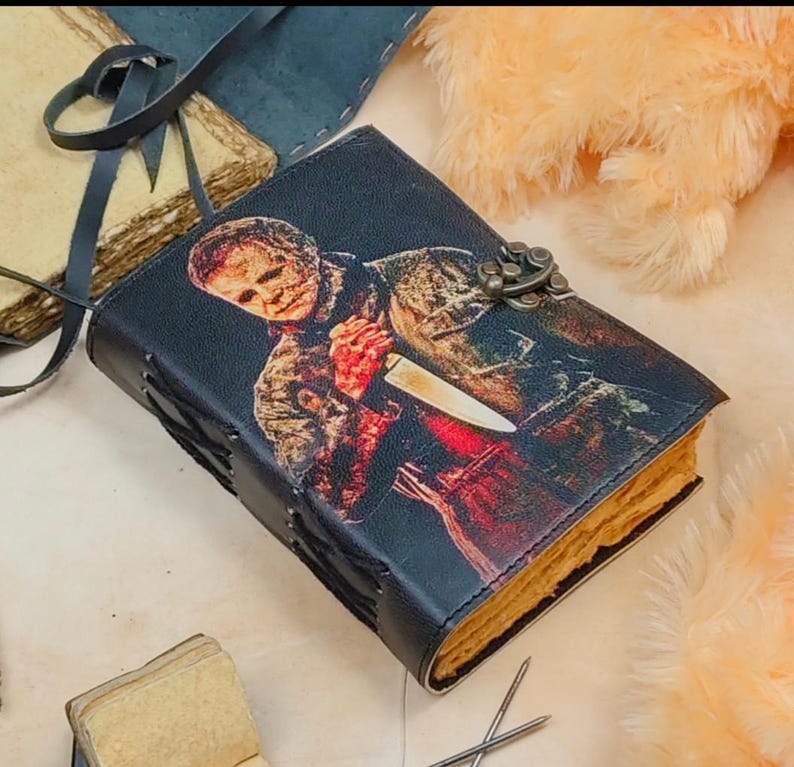 May include: A black leather-bound journal with a graphic of a horror movie character holding a knife. The journal has a metal clasp and is decorated with stitching.