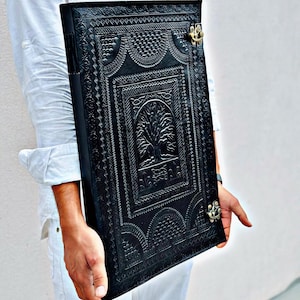 Pode incluir: Di&aacute;rio de couro preto com um design de &aacute;rvore em relevo e fecho de metal decorativo. O di&aacute;rio apresenta padr&otilde;es detalhados e uma superf&iacute;cie texturizada, criando uma est&eacute;tica vintage. Um caderno &uacute;nico para escrever e desenhar.