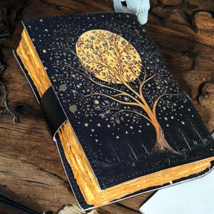 Puede incluir: Un diario de cuero negro con un dise&ntilde;o de &aacute;rbol dorado en la portada. El &aacute;rbol est&aacute; rodeado de estrellas y la luna se representa como un c&iacute;rculo con un &aacute;rbol dorado en su interior. El diario tiene un lomo grueso y est&aacute; cerrado.