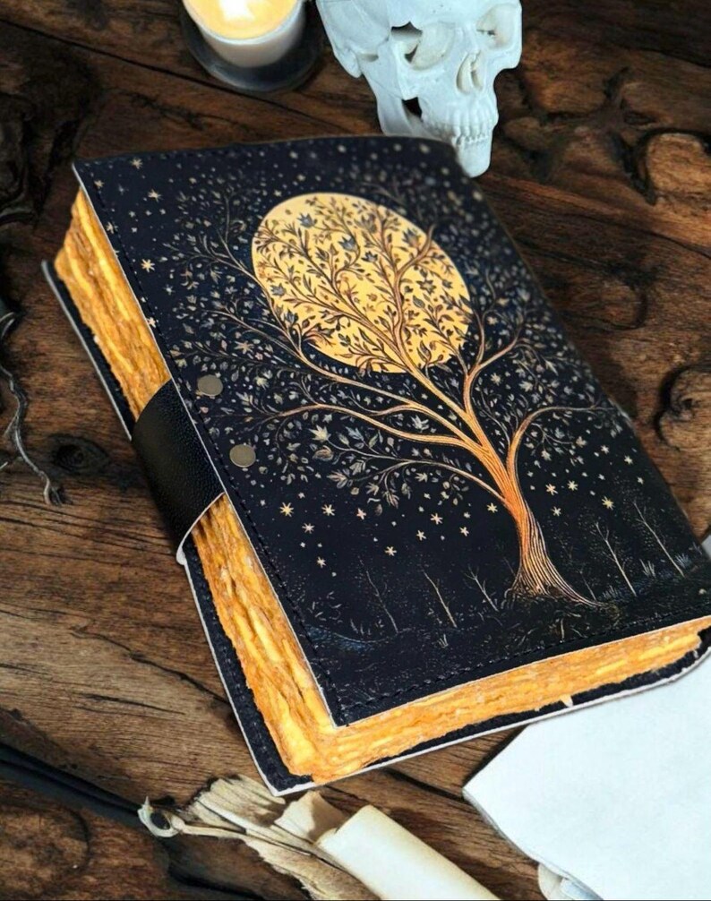 Puede incluir: Un diario de cuero negro con un dise&ntilde;o de &aacute;rbol dorado en la portada. El &aacute;rbol est&aacute; rodeado de estrellas y la luna se representa como un c&iacute;rculo con un &aacute;rbol dorado en su interior. El diario tiene un lomo grueso y est&aacute; cerrado.