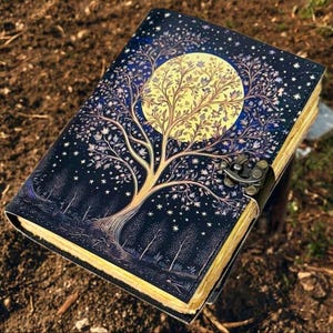 Puede incluir: Un diario de cuero negro con un dise&ntilde;o en relieve dorado y azul de un &aacute;rbol con una luna llena detr&aacute;s. El diario tiene un cierre de metal.