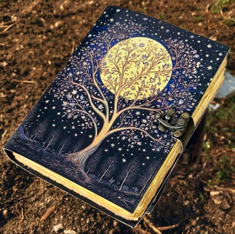 Puede incluir: Un diario de cuero negro con un dise&ntilde;o en relieve dorado y azul de un &aacute;rbol con una luna llena detr&aacute;s. El diario tiene un cierre de metal.