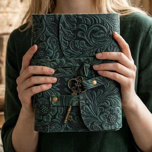 Könnte beinhalten: Ein dunkelgrünes Lederjournal mit geprägtem floralem Design. Das Journal hat einen Riemenverschluss mit einem dekorativen Schlüsselanhänger. Die Hülle ist dunkelgrün und der Stil ist Vintage.